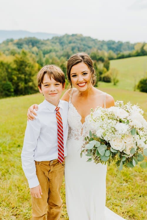 Bride with young boy in a field with rolling hills and trees, holding a white and green bouquet at Moose Hollow Lodge.