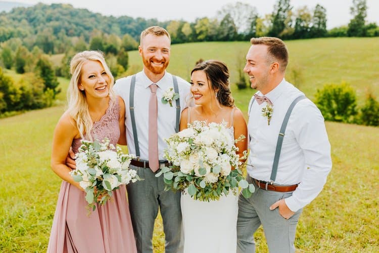 Wedding party of four, two women and two men, standing in a grassy field with green hills and trees in the background.