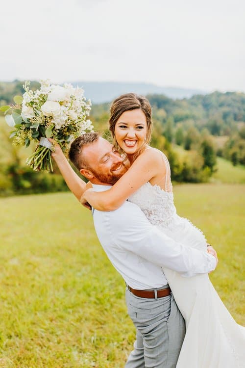 Groom holding bride in a field, her holding a bouquet, with rolling mountains in the background at Moose Hollow Lodge.