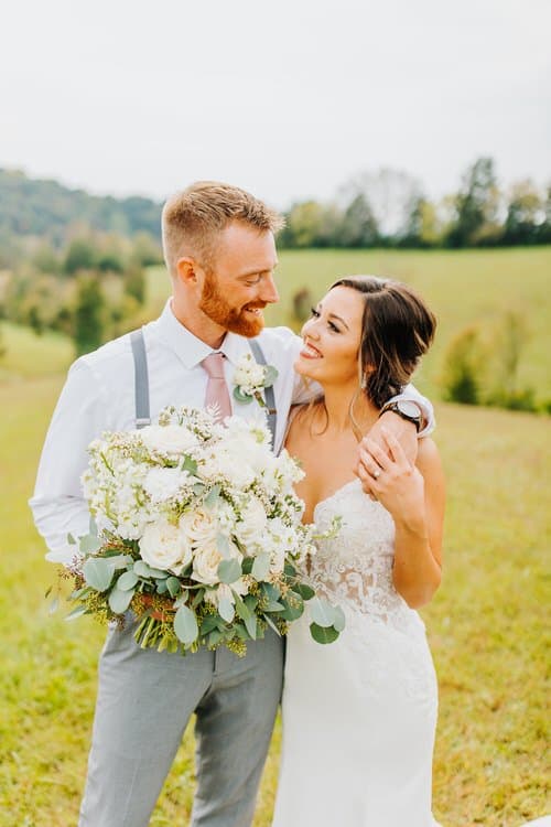 Wedding couple embracing outdoors. Groom in suspenders holds white bouquet, bride in lace dress in grassy field with...