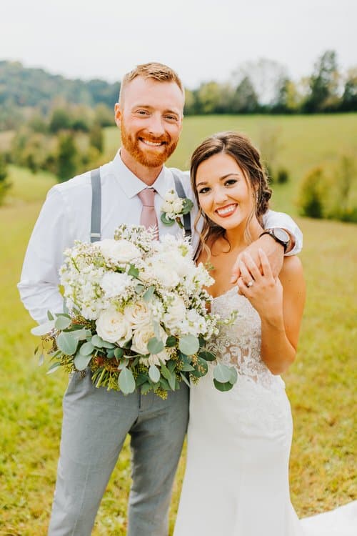 Wedding couple portrait outdoors in a grassy field with hills. Groom in suspenders, bride in lace dress holding white...