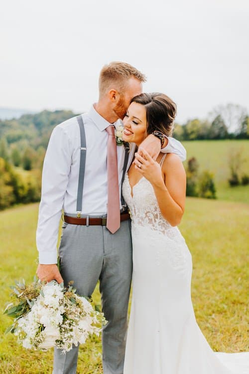 Bride and groom embrace in a grassy field with rolling hills in the background. Groom holds a white floral bouquet.