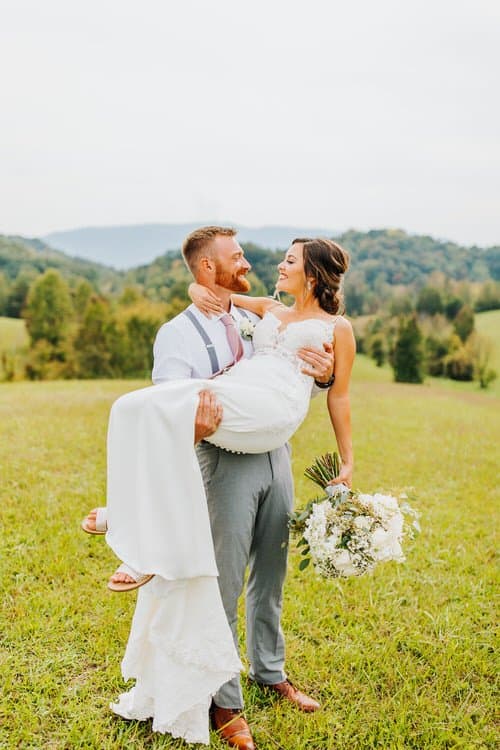 Groom carries bride in a sunny grassy field with distant green hills and trees in the background in the Smoky Mountains.