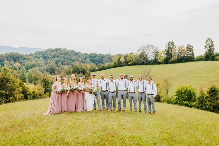 Wedding party, including bride, bridesmaids in long mauve dresses, and groomsmen in suspenders, standing in a field with...