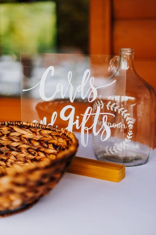 Acrylic 'Cards and Gifts' sign with a wicker basket and glass bottle on a white table. Blurred rustic wooden background.
