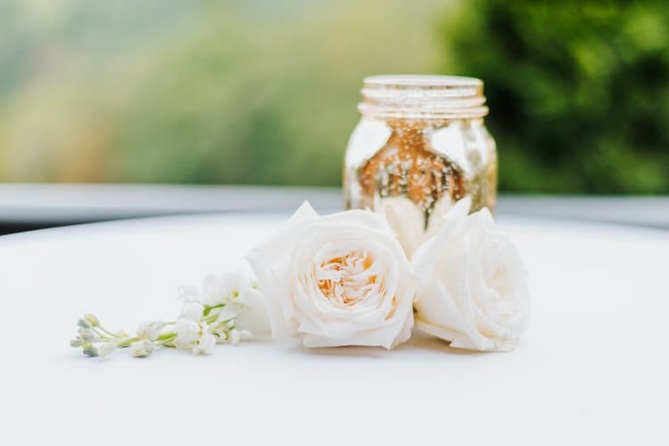 Two white roses, a white floral sprig, and a gold mercury glass jar on a white surface with blurred green background.