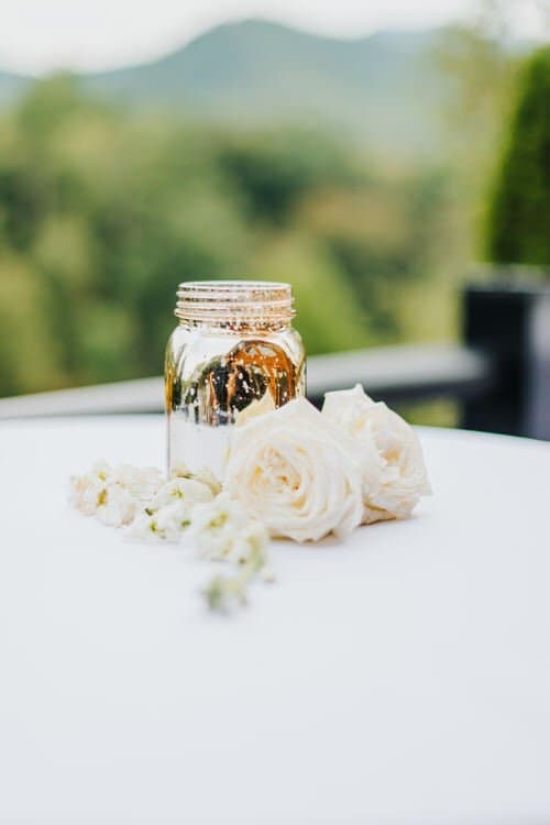 Mercury glass mason jar centerpiece with white roses and filler flowers on a white table, mountains in background.