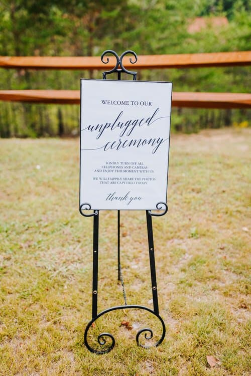 White 'unplugged ceremony' sign on a black easel in grassy outdoor setting with a wooden fence and trees in the background.