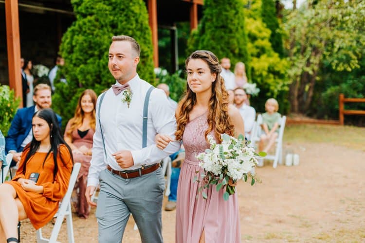 Man in suspenders and woman in pink dress with bouquet walk down aisle at Moose Hollow Lodge outdoor wedding ceremony.