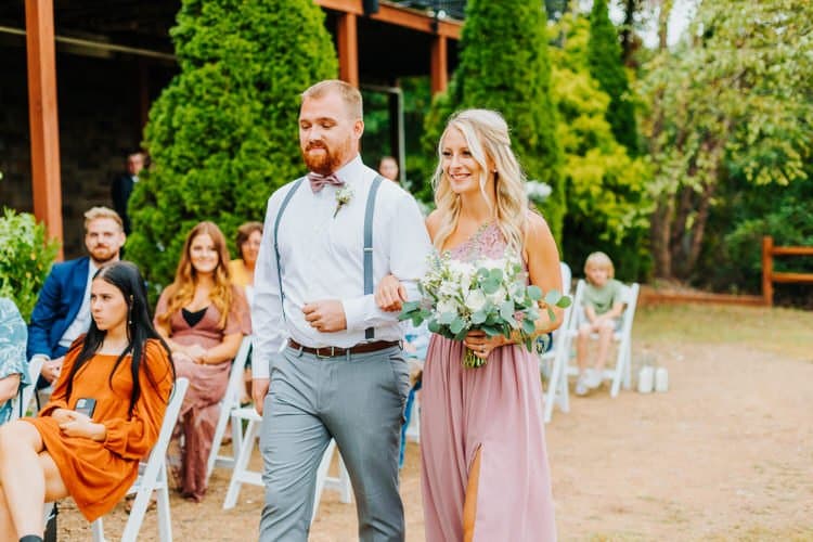 A man in suspenders and a woman in a pink dress holding a bouquet walk down an outdoor aisle past seated guests at Moose...