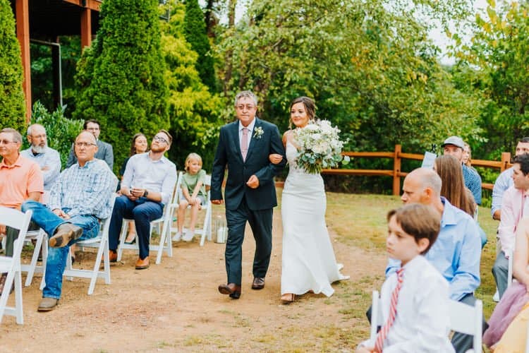 Bride walks down an outdoor aisle with a man in a dark suit past seated guests in white chairs. Green trees in background.