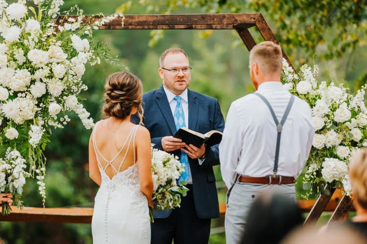 Outdoor wedding ceremony with a bride in a white gown, groom in suspenders, and an officiant under a floral arch.