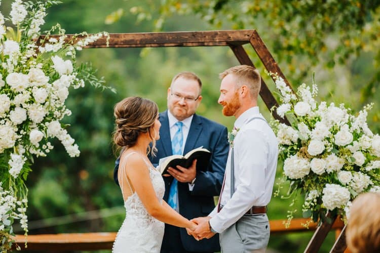 Bride and groom holding hands during an outdoor wedding ceremony at Moose Hollow Lodge. Officiant stands between them...