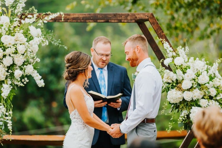 Couple holding hands at an outdoor wedding ceremony with officiant under a wooden arbor decorated with white flowers.