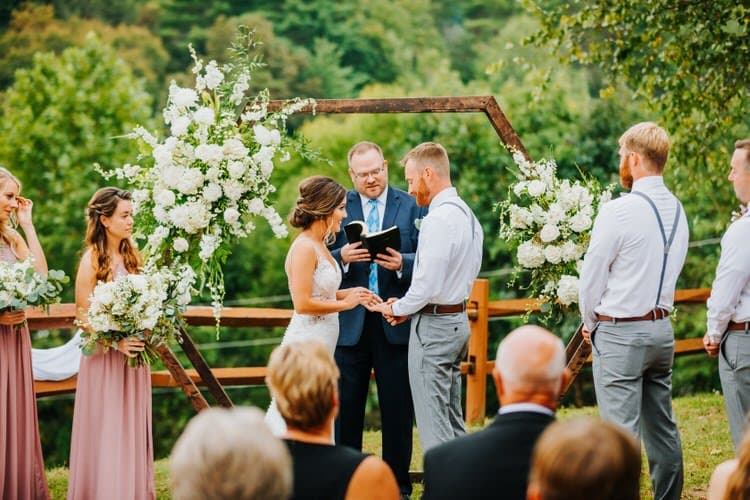 Outdoor wedding ceremony with a couple, officiant, wedding party members, and guests. Green hilly background with a...