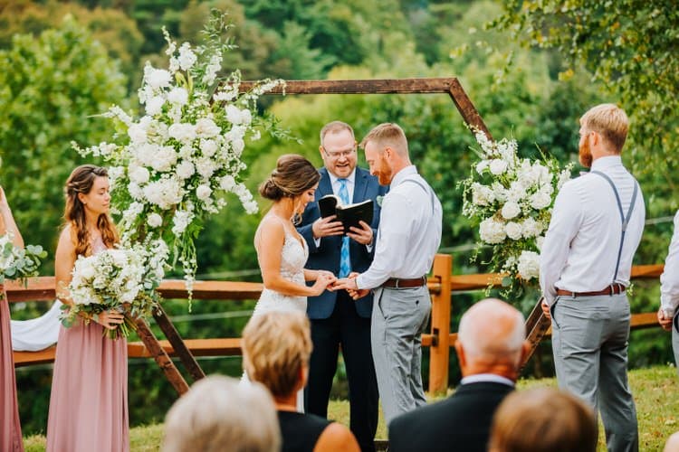 Couple exchanges rings at an outdoor wedding ceremony at Moose Hollow Lodge, officiated under a geometric arch with white...