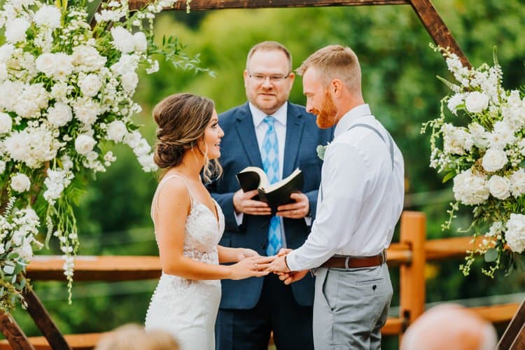 Bride and groom hold hands during an outdoor wedding ceremony at Moose Hollow Lodge. An officiant stands behind them...