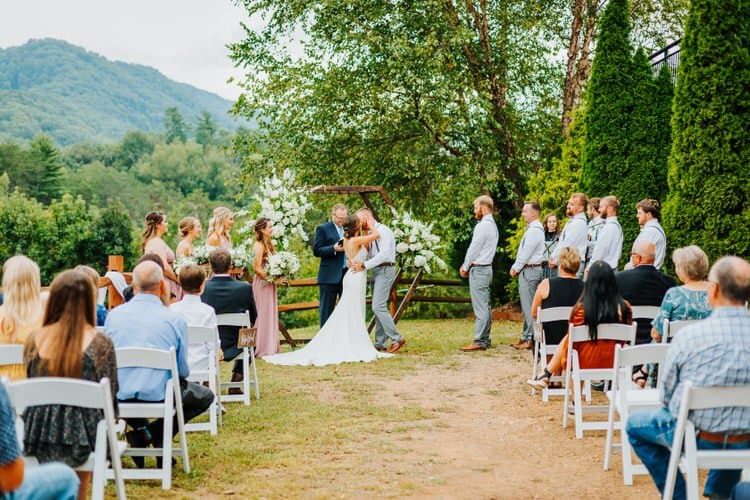 Couple kisses at outdoor wedding ceremony under a floral arch, with bridesmaids, groomsmen, and guests present. Mountains...