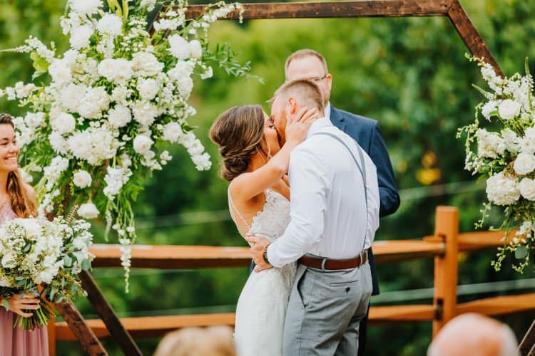 Couple kissing under a white floral-decorated hexagonal arch at an outdoor wedding. Officiant and a bridesmaid are visible.