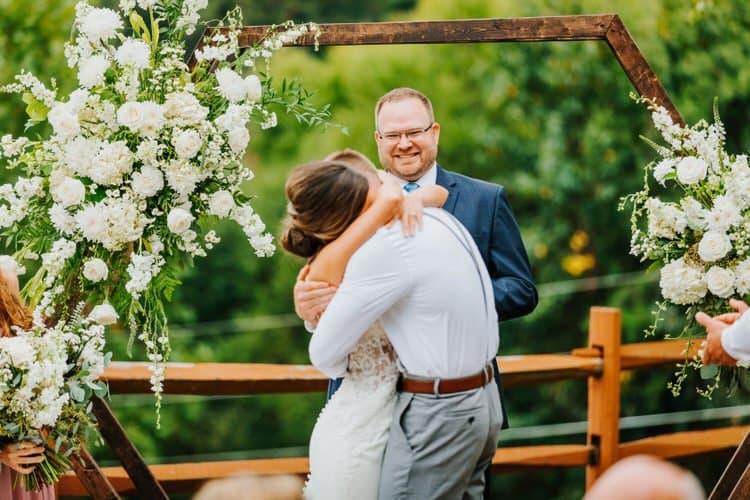 Couple embracing under a wooden hexagonal arch adorned with white flowers at an outdoor wedding ceremony, officiant smiling.