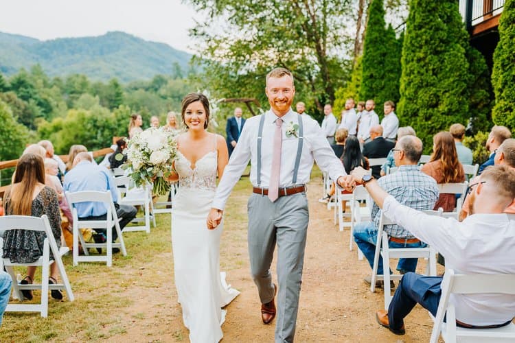 Bride and groom walk hand-in-hand down an outdoor aisle past seated guests, with mountains and trees at Moose Hollow Lodge.