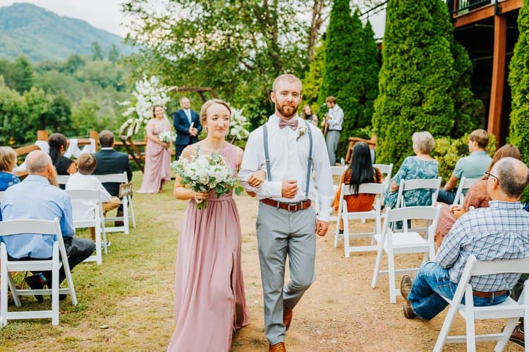 Woman in pink dress and man in white shirt walk arm-in-arm down an aisle past seated guests outdoors at Moose Hollow...