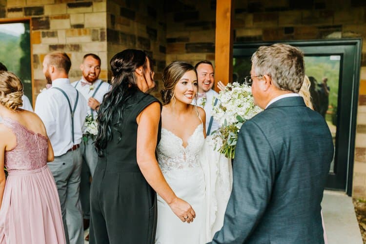 Bride in white gown holding bouquet, smiling at guests on a stone and wood porch at Moose Hollow Lodge. Groomsmen in...