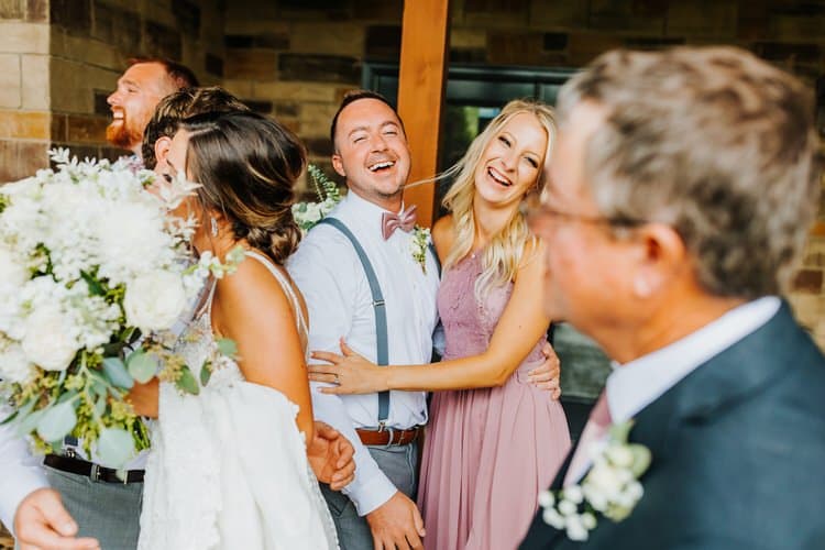 Wedding guests and a couple laughing outdoors at a stone and wood lodge. A bride holds a white bouquet.