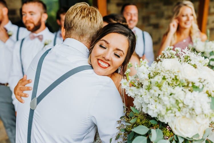 A woman holding a white and green floral bouquet smiles with closed eyes while embracing a man in a white shirt and grey...