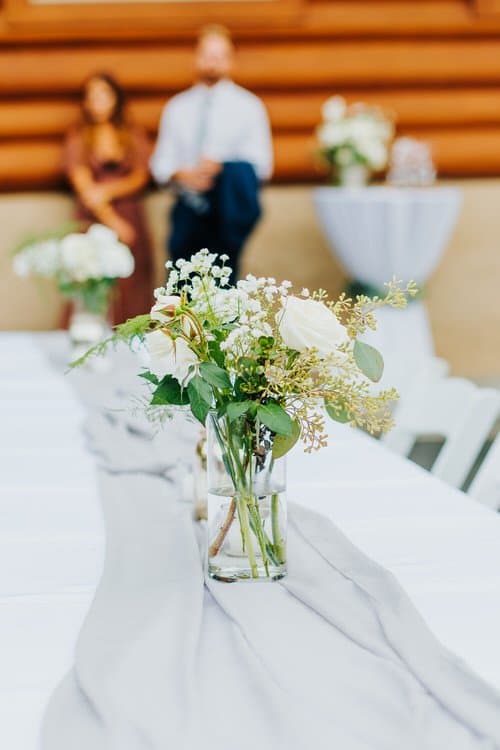 White roses and greenery centerpieces on a reception table with a gray runner, blurred guests and log cabin background.