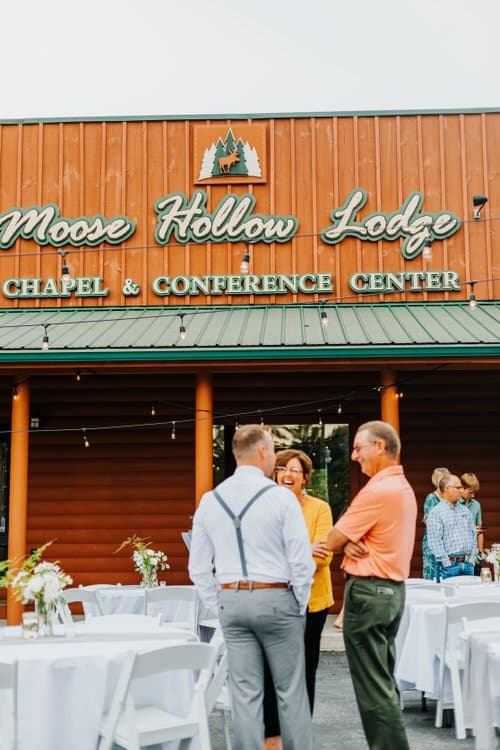 Guests mingle at outdoor reception tables in front of Moose Hollow Lodge, a log cabin venue with its sign overhead.