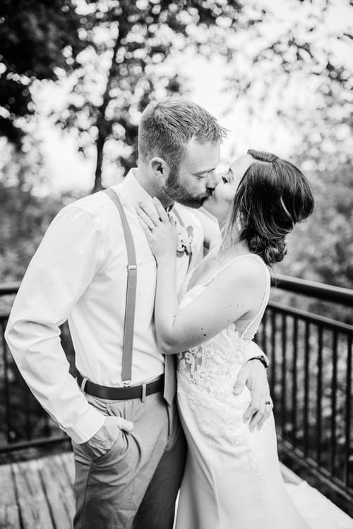 Black and white photo of a wedding couple kissing outdoors on a wooden deck with a metal railing, trees in background.