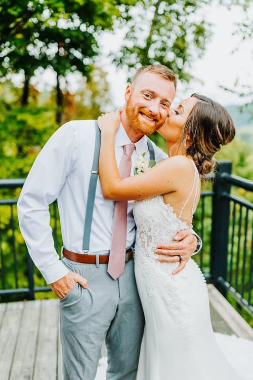 Couple portrait on a wooden deck with a metal railing. Groom in a white shirt and gray suspenders, bride in a white lace...