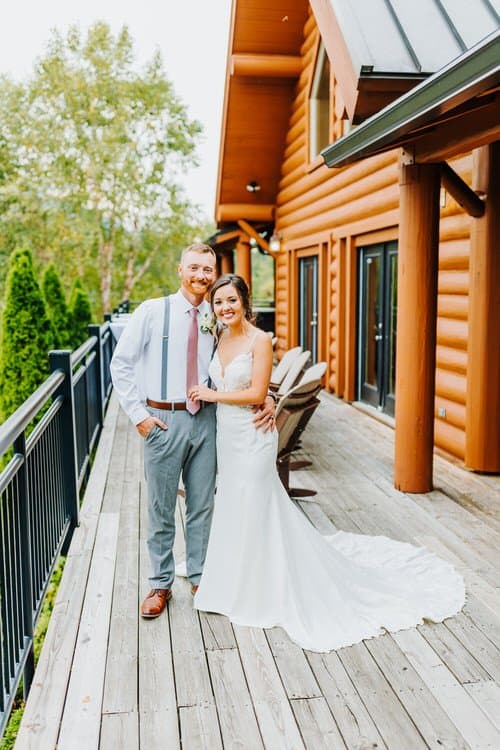 Wedding couple stands smiling on a wooden deck outside a log cabin at Moose Hollow Lodge, surrounded by greenery.