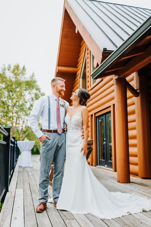 Bride and groom on a wooden deck at Moose Hollow Lodge. A large log cabin with a dark roof is behind them, with trees and...