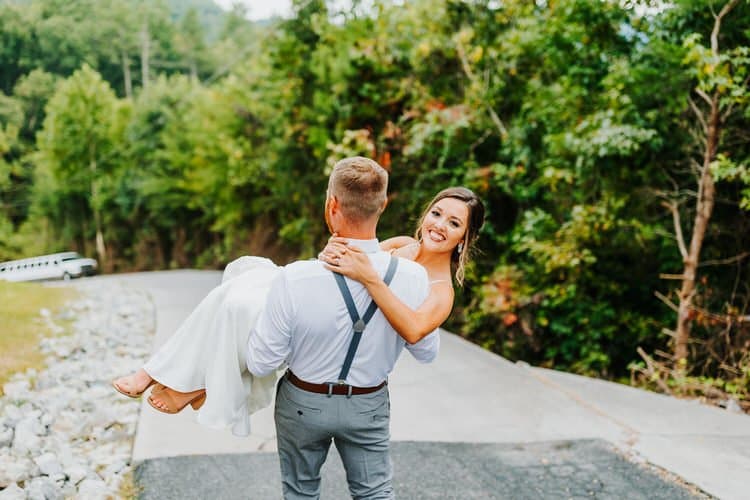 Groom carries bride outdoors on a paved path. Lush green trees and a white limousine are visible in the background at...