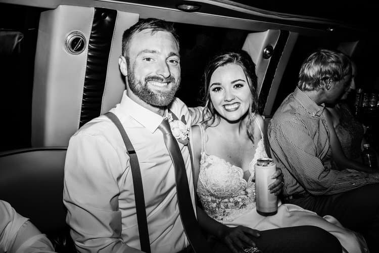 Groom and bride smiling in a limousine, she holds a tumbler. Black and white photo with another passenger.