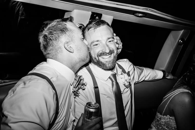 Two men in wedding attire share a kiss inside a vehicle. One man holds a can and smiles. Black and white image.