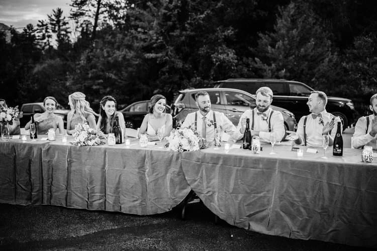Wedding party seated at a long outdoor table with florals, candles, and champagne. Trees and cars in background.