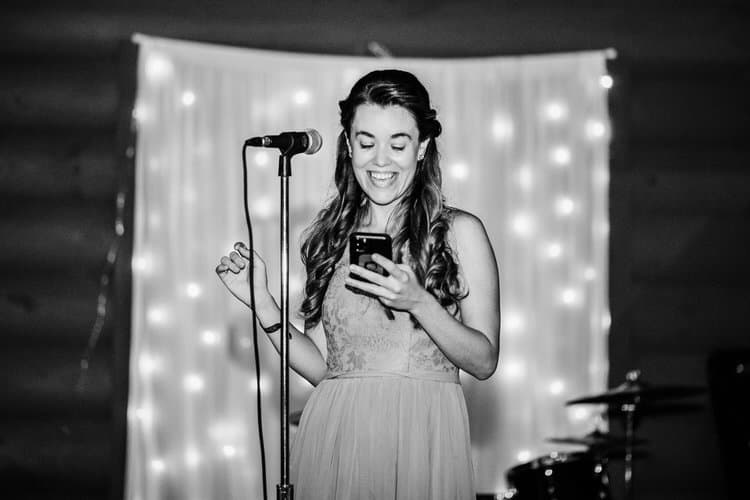 A woman with long hair holds a smartphone and stands by a microphone, laughing. White drapes with string lights are...