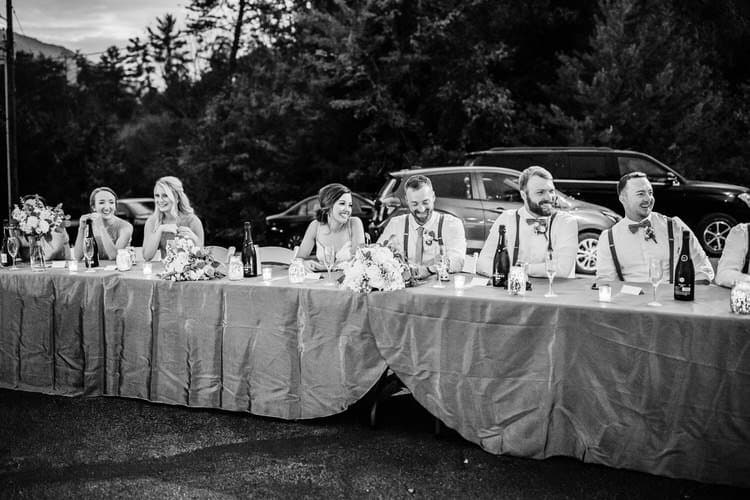 Wedding party seated at an outdoor head table, smiling. Forested background with mountains and parked cars. Evening lighting.