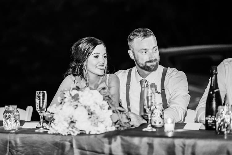 Black and white photo of a smiling woman and bearded man seated at a wedding table with a floral centerpiece and drinks.