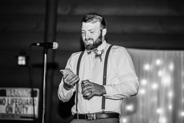 Man in suspenders and bow tie with boutonnière holds phone and drink, standing by a microphone at Moose Hollow Lodge.