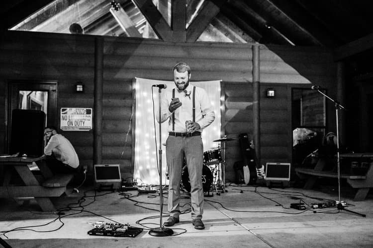 Man in bow tie and suspenders holding phone and drink at microphone, inside Moose Hollow Lodge cabin venue with band...