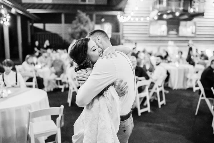 Couple dancing at a wedding reception, black and white photo. Guests seated at tables with string lights overhead.