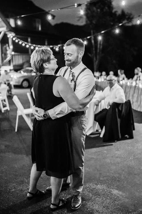 A woman in a dark dress dances with a man in suspenders and a white shirt under string lights at an outdoor evening event.
