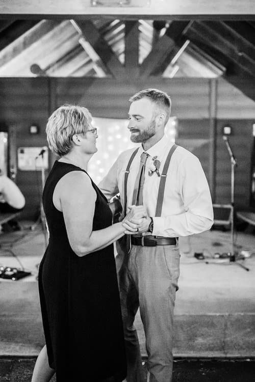 Groom in suspenders dancing with a woman in a black dress, under wooden beams at Moose Hollow Lodge wedding.
