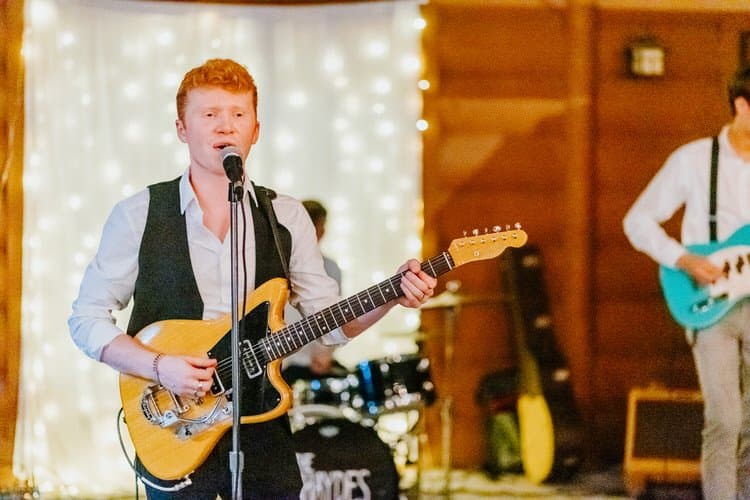 Musician with red hair sings into a microphone, playing an electric guitar. String lights and wooden walls in background.
