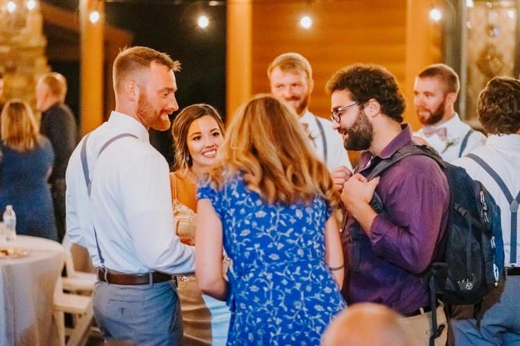 Group of wedding guests, including a smiling woman in a light dress, socializing at an evening reception with string lights.