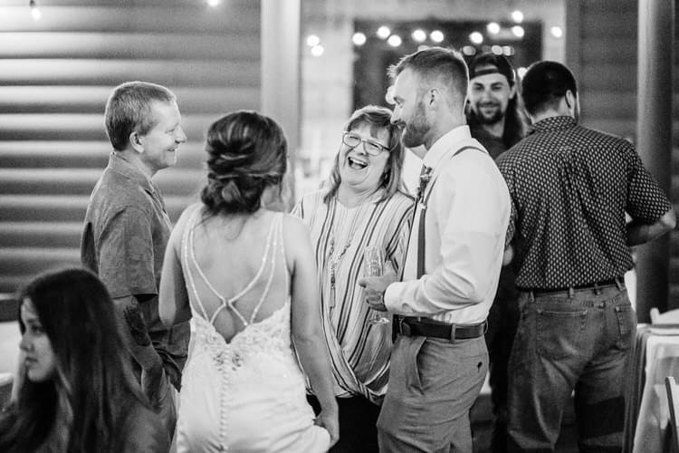 Wedding guests mingle in black and white, including bride and laughing woman. Log cabin wall, string lights at Moose...